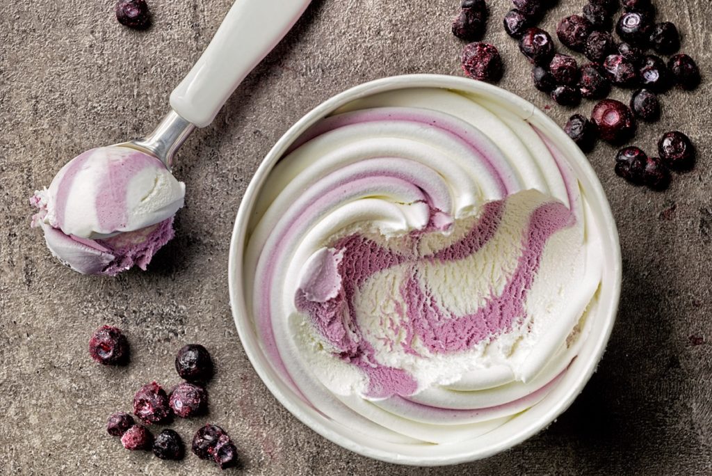 Înghețată de vanilie cu afine în bol alb, alături de o lingură cu înghețată și afine proaspete