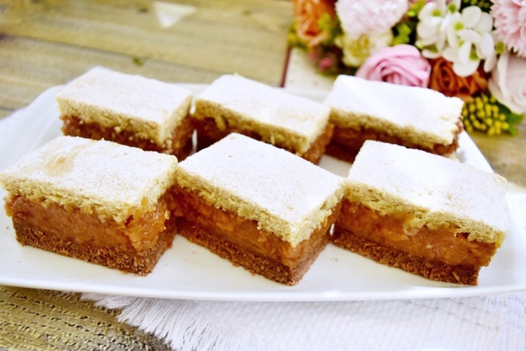 Porții de prăjitură cu mere și aluat bicolor, pe un platou alb, alături de un buchet de flori