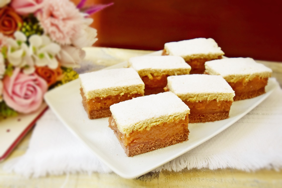 Porții de prăjitură cu mere și aluat bicolor, pe un platou alb, alături de un buchet de flori