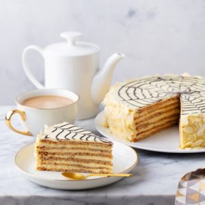 Porție de tort Esterhazy pe o farfurie albă, alături de tortul secționat și recipiente albe cu cafea