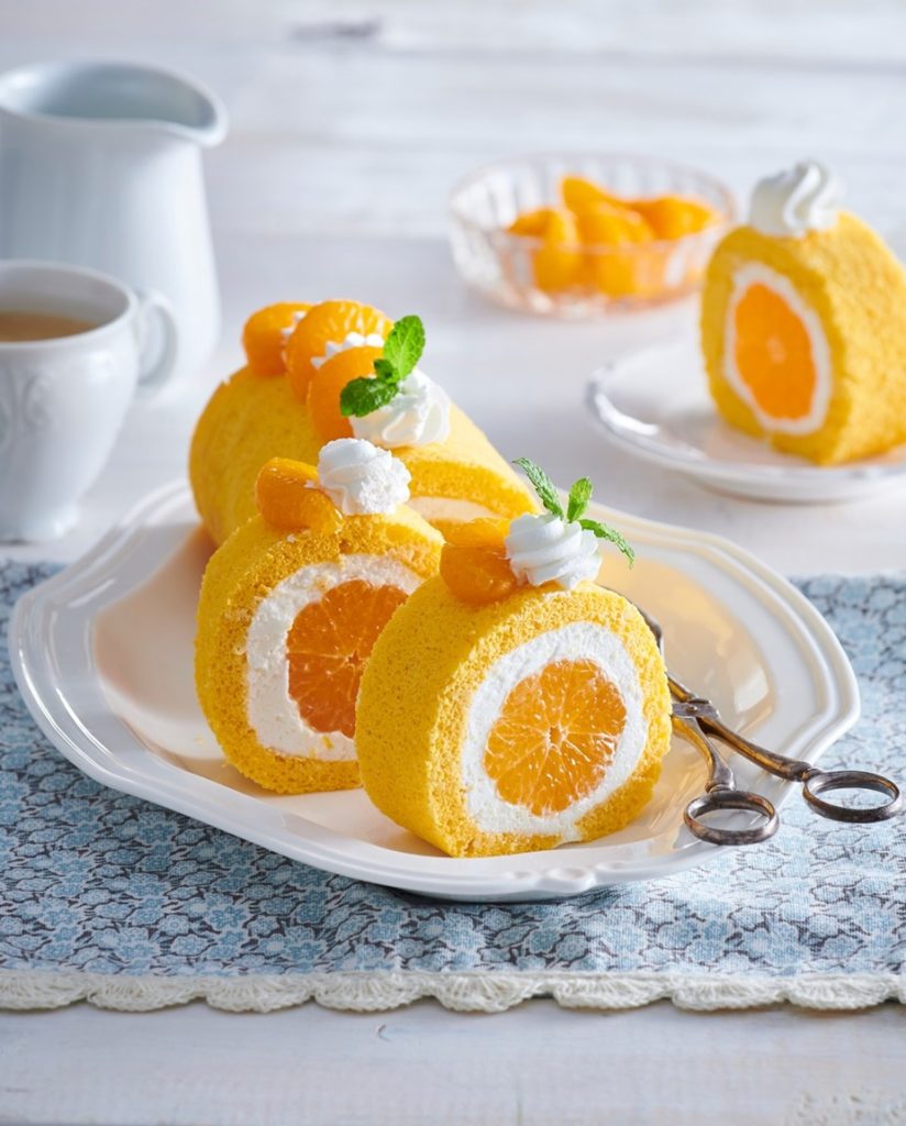 Ruladă cu portocale și cremă de mascarpone porționată pe o farfurie albă, alături de un ceainic