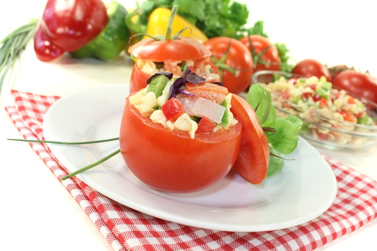 Două roșii umplute pe o farfurie, alături de legume proaspete și un bol cu salată de paste