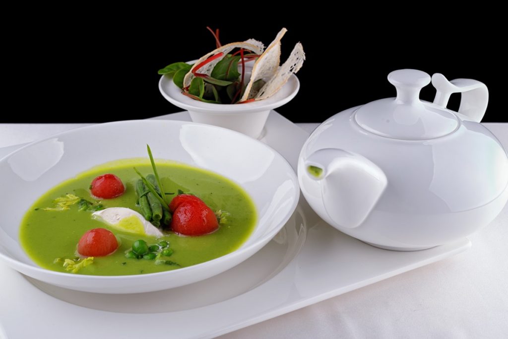 Porție de Supă cremă de mazăre cu roșii coapte în bol alb, alături de un recipient alb cu supă și un bol cu parmezan și verdeață, pe un platou alb