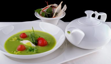 Porție de Supă cremă de mazăre cu roșii coapte în bol alb, alături de un recipient alb cu supă și un bol cu parmezan și verdeață, pe un platou alb
