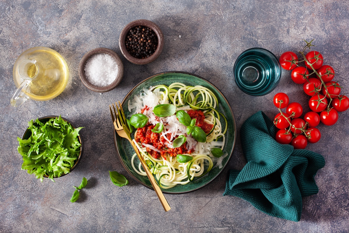 Bol cu Spaghete de dovlecei cu roșii coapte și parmezan alături de roșii proaspete, un bol cu salată verde și recipiente cu condimente