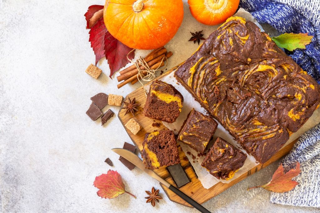 Prăjitură cu ciocolată marmorată cu dovleac plăcintar, porționată pe un platou de lemn, alături de doi dovleci, ciocolată și frunze de toamnă