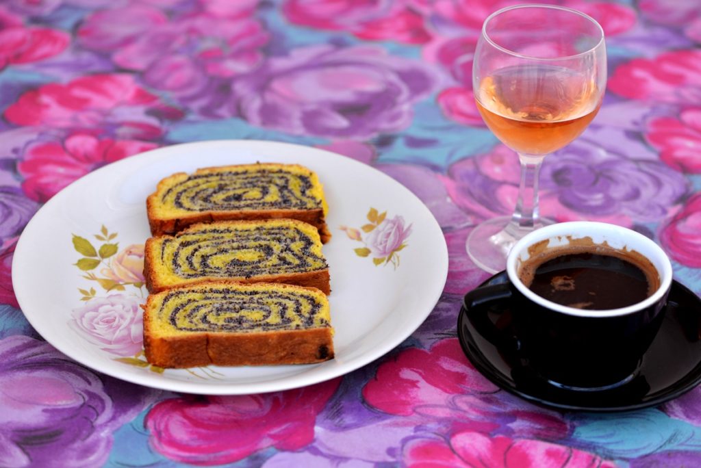 Trei felii de ruladă pe o farfurie albă cu flori, alături de o ceașcă cu cafea și un pahar cu vin