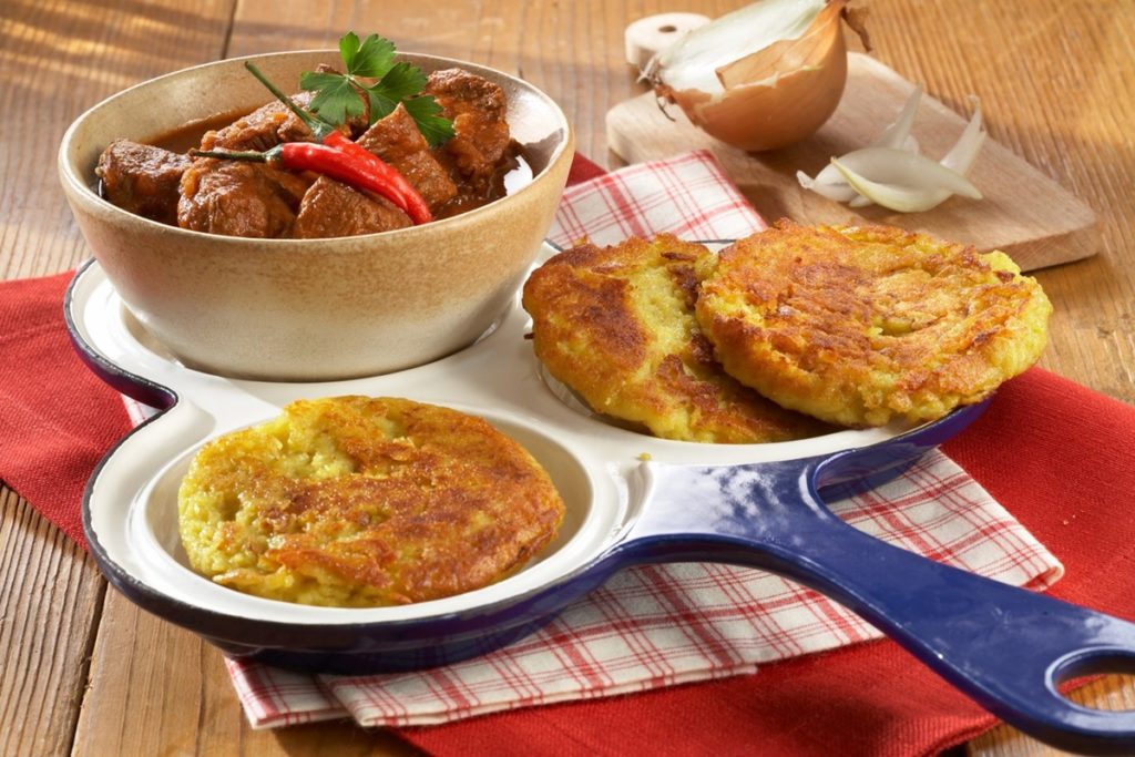 Trei mini omlete cu cartofi rași pe un platou alb cu albastru, alături de un bol cu carne în sos de roșii și o ceapă tăiată