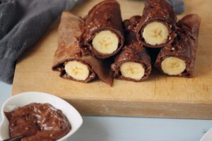 Clătite cu aluat de ciocolată umplute cu cremă și banane pe un suport de lemn, alături de un bol cu cremă de ciocolată