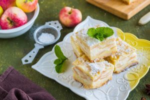 Prăjitură cu mere și brânză porționată pe o farfurie albă și pe un suport de lemn, decorată cu frunze de mentă, alături de un bol cu mere și o sită cu zahăr