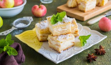Prăjitură cu brânză și mere porționată pe o farfurie albă și pe un suport de lemn, decorată cu frunze de mentă, alături de un bol cu mere