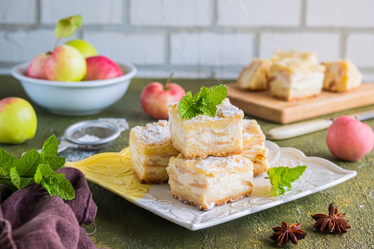 Prăjitură cu mere și brânză porționată pe o farfurie albă și pe un suport de lemn, decorată cu frunze de mentă, alături de un bol cu mere