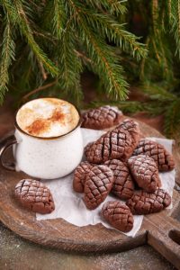 Biscuiți conuri de brad cu cacao pe un tocător de lemn rotund, alături de o cană cu caffe latte și crengi de brad
