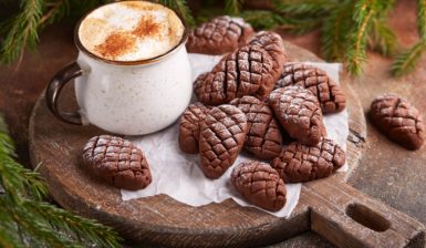 Biscuiți conuri de brad cu cacao pe un tocător de lemn rotund, alături de o cană cu caffe latte și crengi de brad