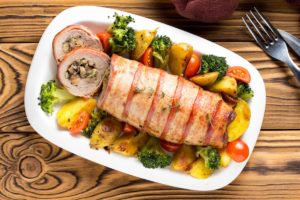 Ruladă de porc cu ciuperci și cașcaval în felii de bacon, pe un platou alb cu cartofi, broccoli și roșii