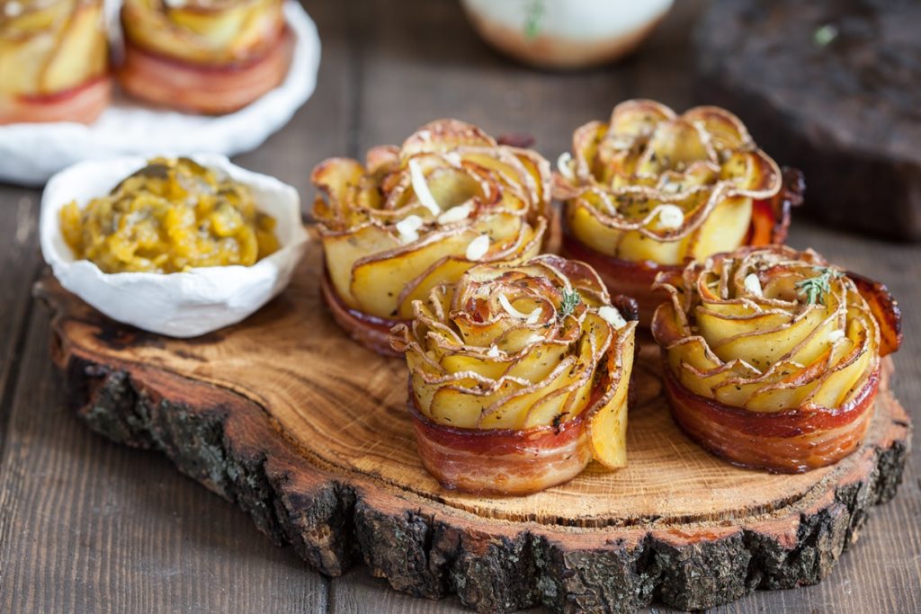 Patru trandafiri crocanți din cartofi cu bacon așezați pe un suport rustic de lemn, alături de un bol cu salată