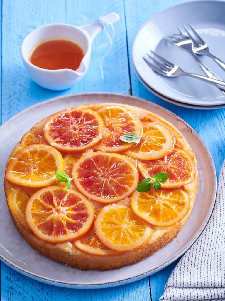 Prăjitură răsturnată cu portocale pe un platou alb, alături de o ceașcă cu sirop, tacâmuri și farfurii