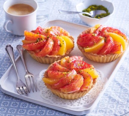 Trei mini tarte cu cremă și portocale pe un platou cu trei furculițe, o ceașcă de cafea și un bol cu fistic mărunțit