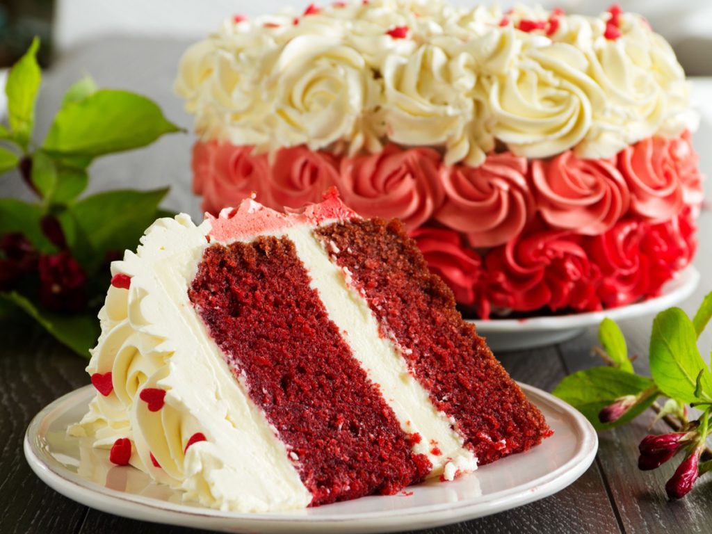 Porție de Red Velvet Cake pe farfurie, alături de platoul cu tort