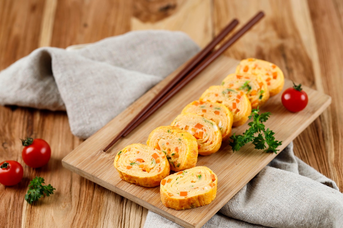 Ruladă din omletă cu morcovi și brânză porționată pe un tocător de lemn, alături de două bețe chinezești
