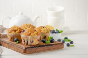 Muffins cu lămâie și afine pe un suport de lemn, alături de un ceainic alb și două cești albe și un bol cu afine