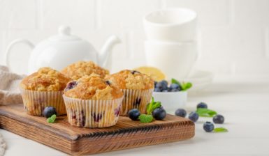 Muffins cu lămâie și afine pe un suport de lemn, alături de un ceainic alb și două cești albe și un bol cu afine