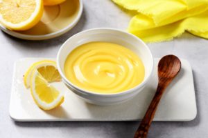 Lemon curd în bol alb, alături de o lingură de lemn și felii de lămâie