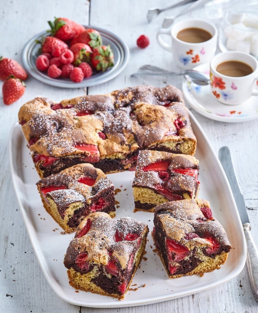 Prăjitură bicoloră cu căpșuni și zmeură porționată pe un platou, alături de două cești cu cafea și o farfurie cu căpșuni