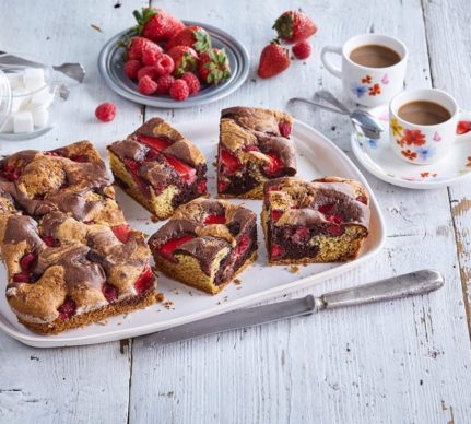 Prăjitură bicoloră cu căpșuni și zmeură porționată pe un platou, alături de două cești cu cafea și o farfurie cu căpșuni