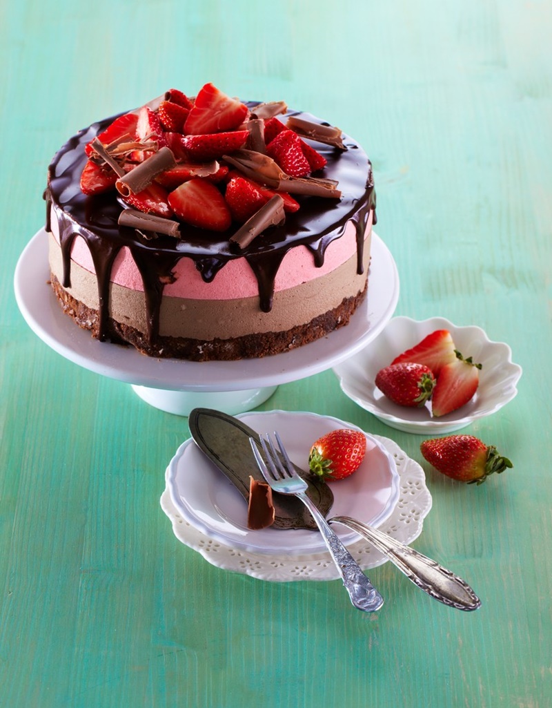 Tort cu două tipuri de mousse și glazură de ciocolată pe un platou cu picior, alături de farfurii și tacâmuri și un bol cu căpșuni