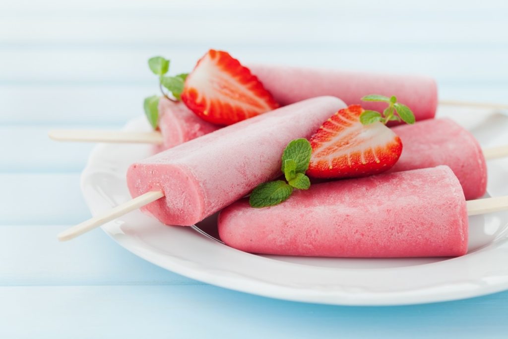 Cinci înghețate pe bețe așezate pe o farfurie și decorate cu căpșuni și mentă