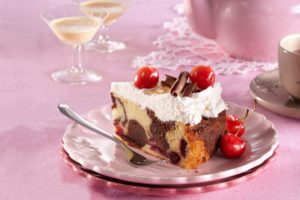 Porție de tort cu blat bicolor și cireșe, cremă de frișcă și decor din ciocolată și cireșe, pe o farfurie cu furculiță, alături de un ceainic și două pahare cu lichior