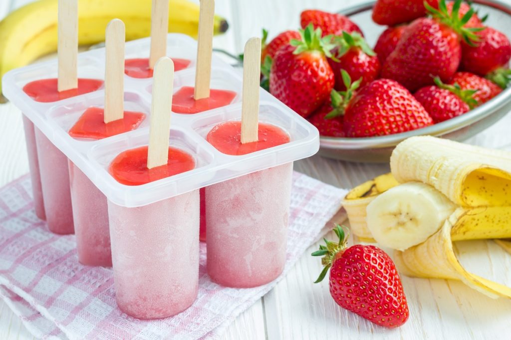 Formă de plastic cu șase înghețate de căpșuni cu banane pe bețe, alături de căpșuni și banane proaspete