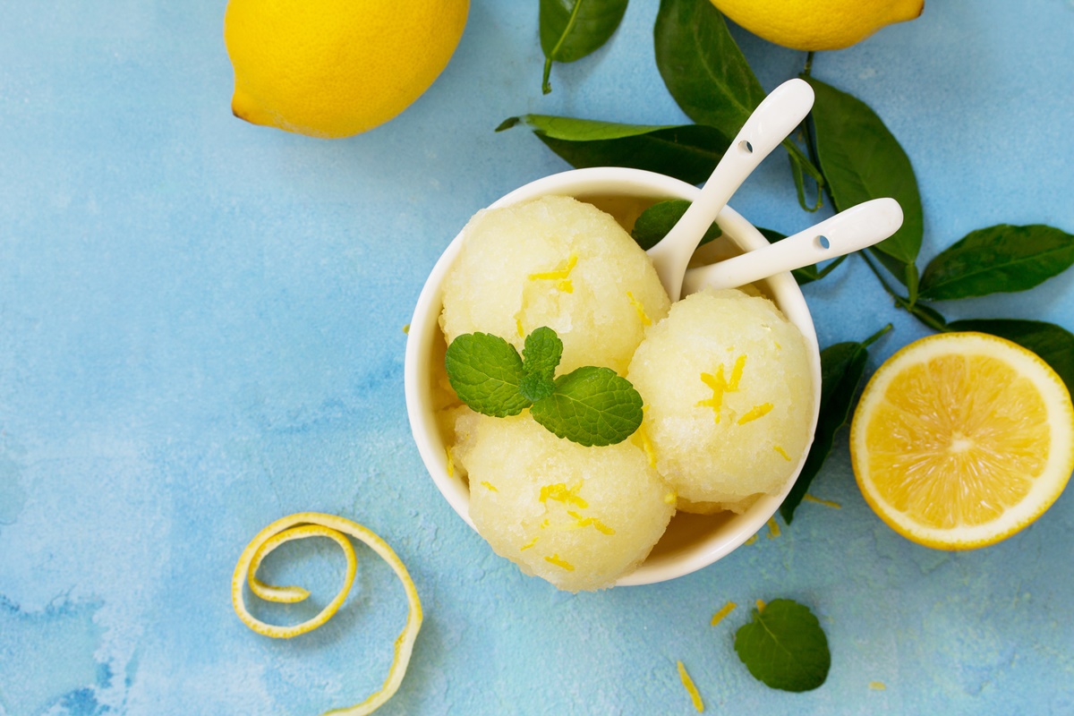Înghețată cu aromă intensă de lămâie în bol alb cu lingurile, alături de lămâi cu frunze