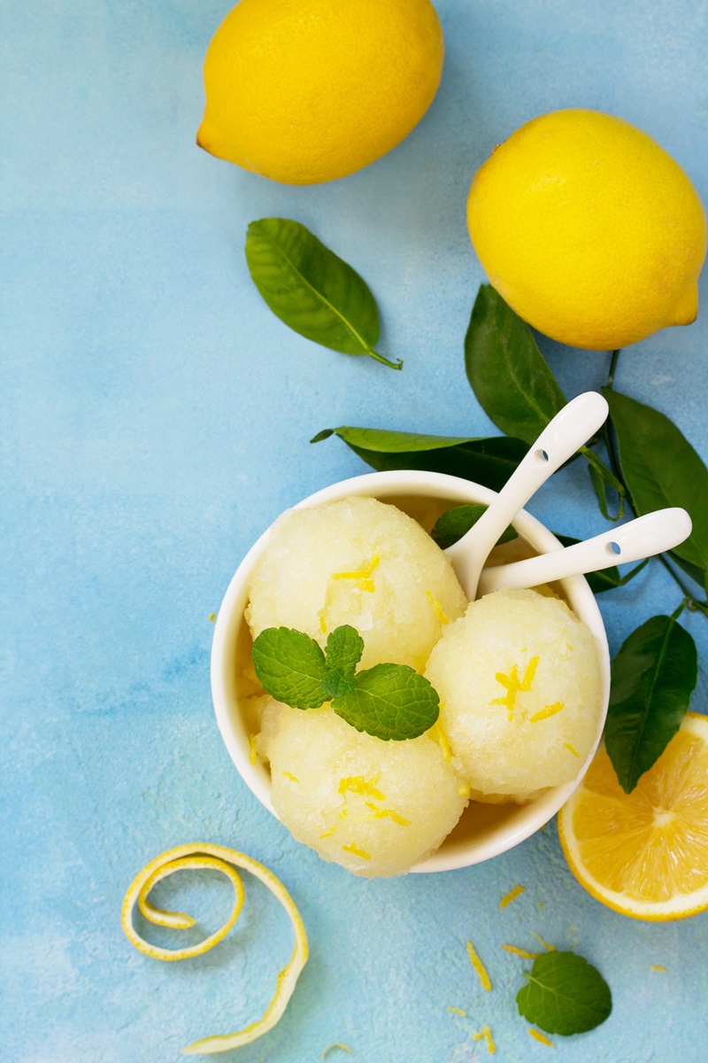 Înghețată cu aromă intensă de lămâie în bol alb cu lingurile, alături de lămâi cu frunze