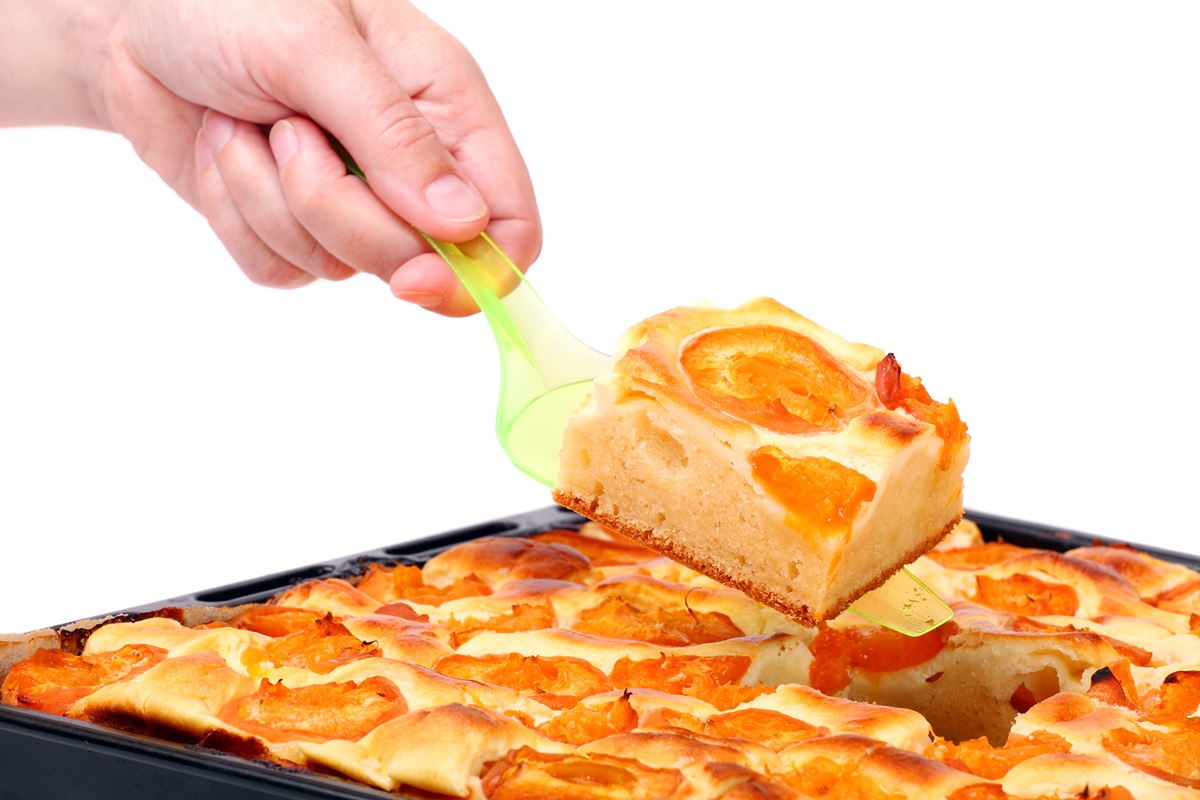 Femeie ridicând cu spatula o porție de prăjitură cu brânză și caise din tavă