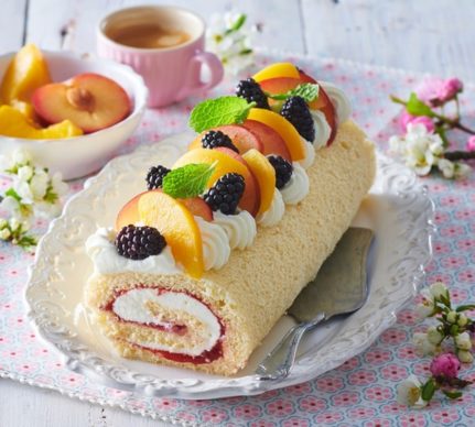 Ruladă cu mascarpone și fructe pe un platou cu spatulă de tort, alături de o ceașcă cu cafea și un bol cu fructe