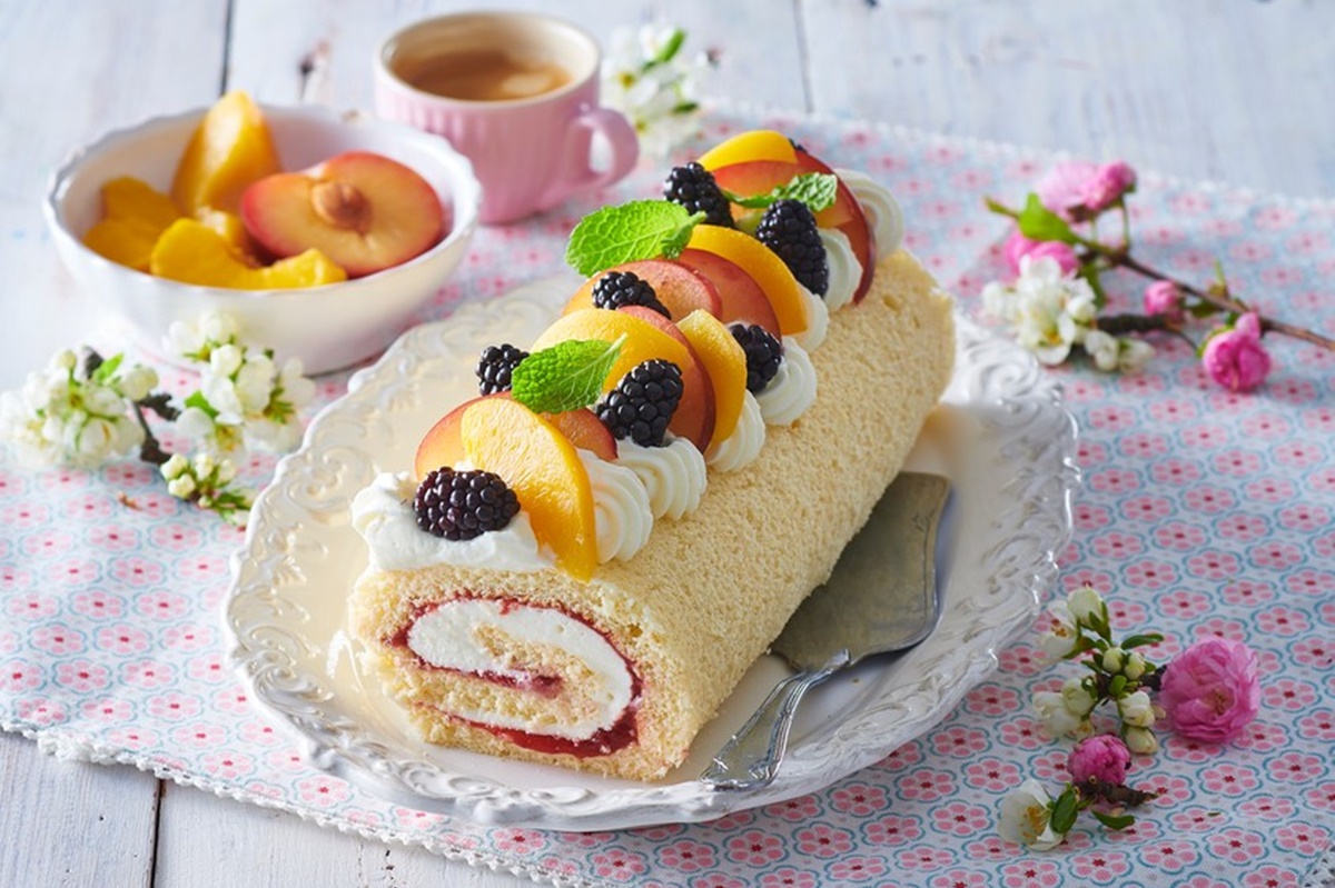 Ruladă cu mascarpone și fructe pe un platou cu spatulă de tort, alături de o ceașcă cu cafea și un bol cu fructe
