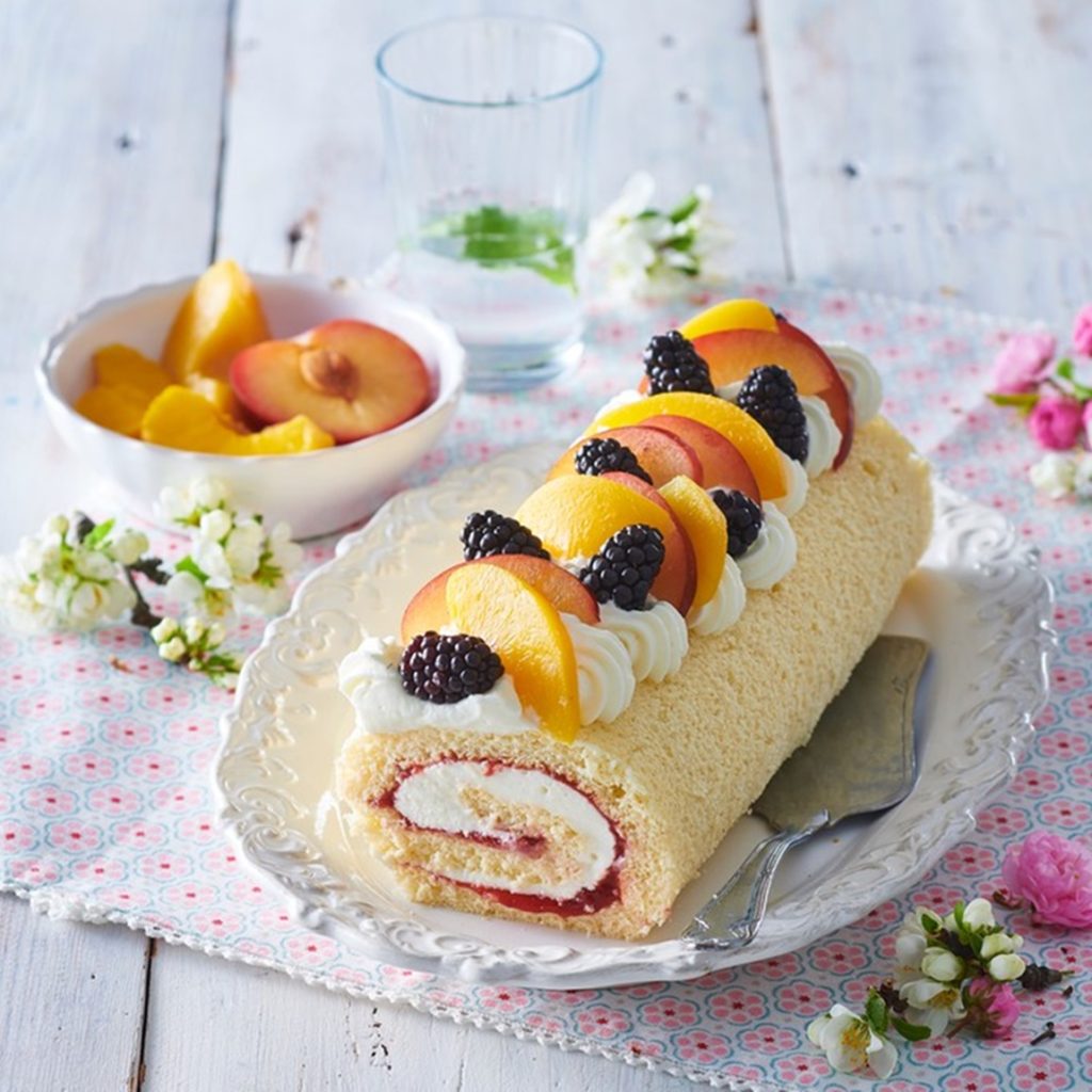 Ruladă cu mascarpone și fructe pe un platou cu spatulă de tort, alături de un pahar și un bol cu fructe