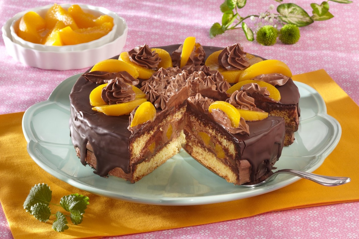 Tort cu ciocolată și piersici din compot porționat pe un platou rotund, alături de un bol cu piersici