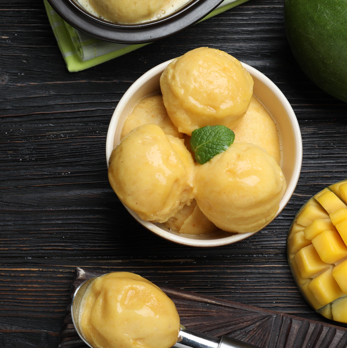 Înghețată de mango în bol și în linguriță, alături de un fruct mango secționat în cuburi