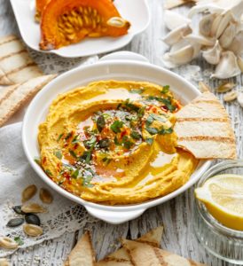 Hummus cu dovleac copt în bol alb, alături de lipii de casă, usturoi, o jumătate de lămâie într-un bol de sticlă și două felii de dovleac cu semințe