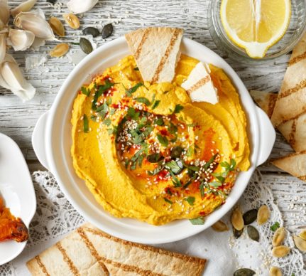Hummus cu dovleac copt în bol alb, alături de lipii de casă, usturoi, o jumătate de lămâie într-un bol de sticlă și două felii de dovleac cu semințe