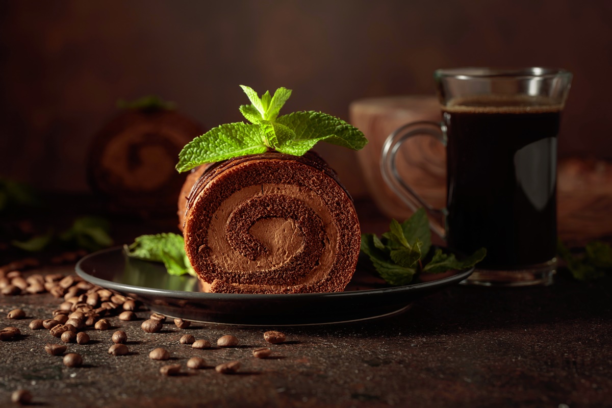 Porție de ruladă cu cremă de cafea și mascarpone, decorată cu frunze de mentă, alături de o ceașcă cu cafea