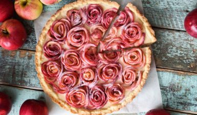 Tartă cu trandafiri din mere și cremă de vanilie porționată pe hârtie de copt, alături de mere proaspete