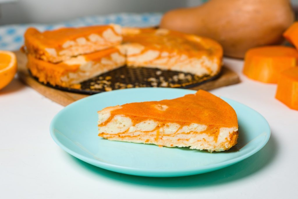 Porție de cheesecake zebră cu dovleac și portocale pe o farfurie, alături de tava cu cheesecake