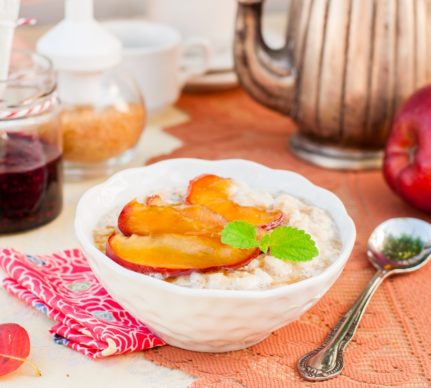 Porție de porridge cu mere în sos caramel în bol alb, alături de un set de ceai și o lingură