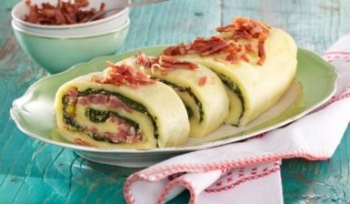 Ruladă de cartofi cu spanac și bacon porționată pe un platou și decorată cu bacon, alături de un bol cu bucăți de bacon