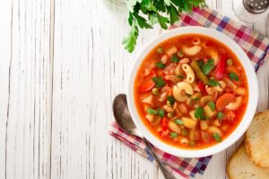 Porție de minestrone în bol alb cu lingură, felii de pâine, o solniță și pătrunjel verde