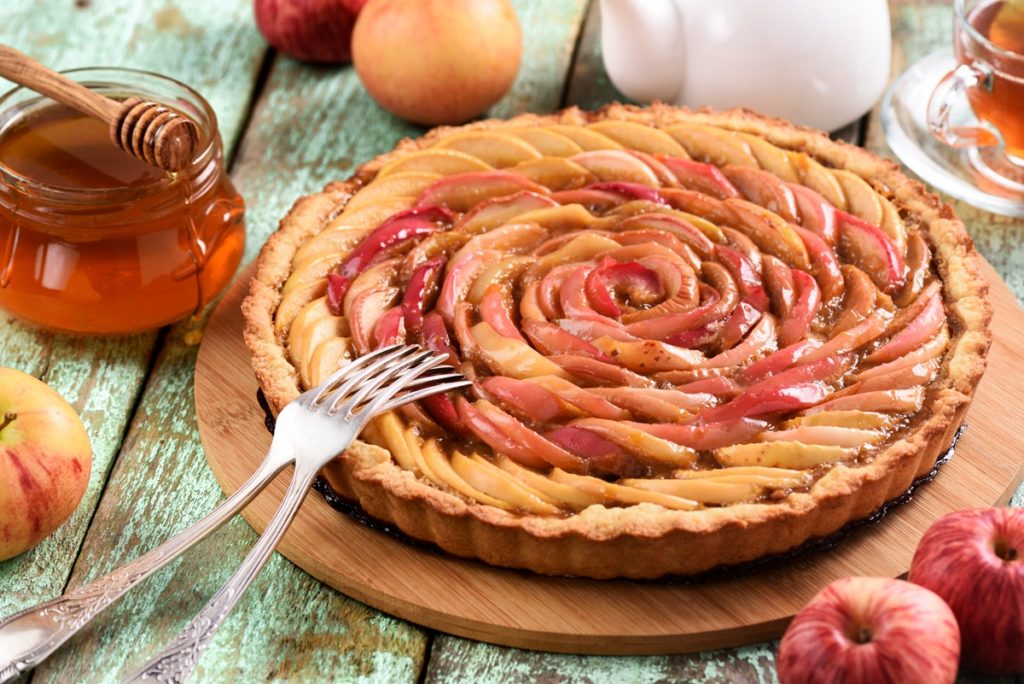 Tartă cu mere sub formă de trandafir pe un platou de lemn, alături de două furculițe, un borcan cu miere și ustensilă de lemn, un ceainic alb și o ceașcă de ceai, toate înconjurate de mere